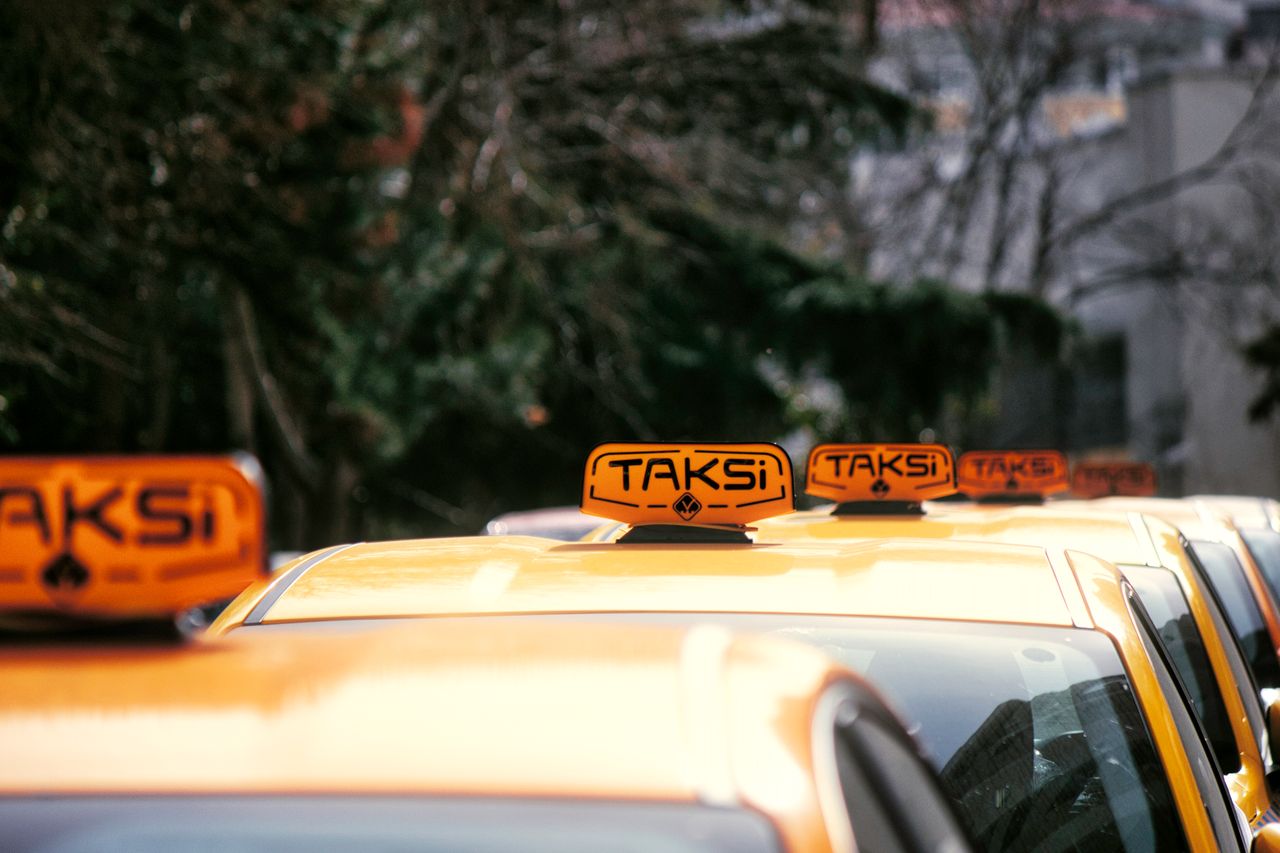 A row of yellow taxis with 'Taksi' signs parked on a city street in daylight.