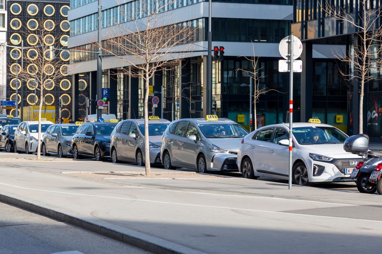 A lineup of taxis on an urban street with modern buildings in the background.